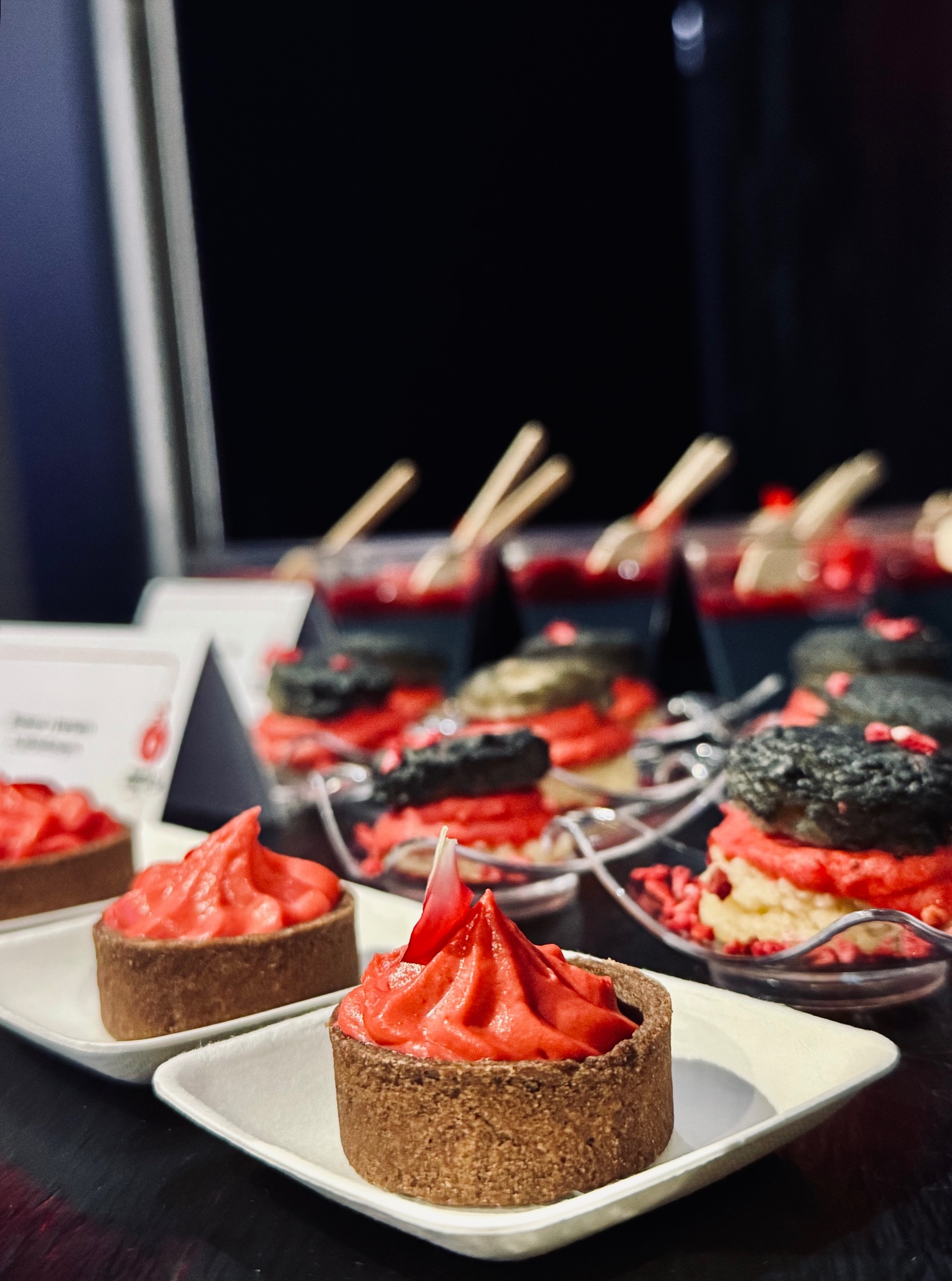 Artystyczna prezentacja czerwonych tartaletek i deserów w szklankach na ciemnym tle, prawdopodobnie na evencie kulinarnym. Unikalne ujęcie z bliska.