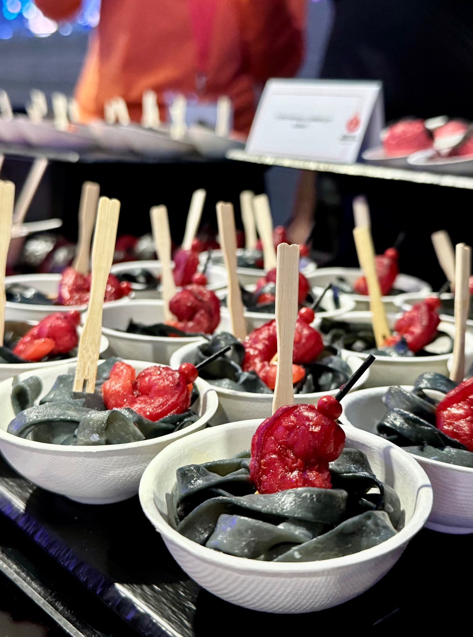 Czarny makaron z czerwonymi dodatkami w białych miseczkach, podany na czarnym stole z drewnianymi widelczykami. Catering na evencie, oświetlenie sceniczne.