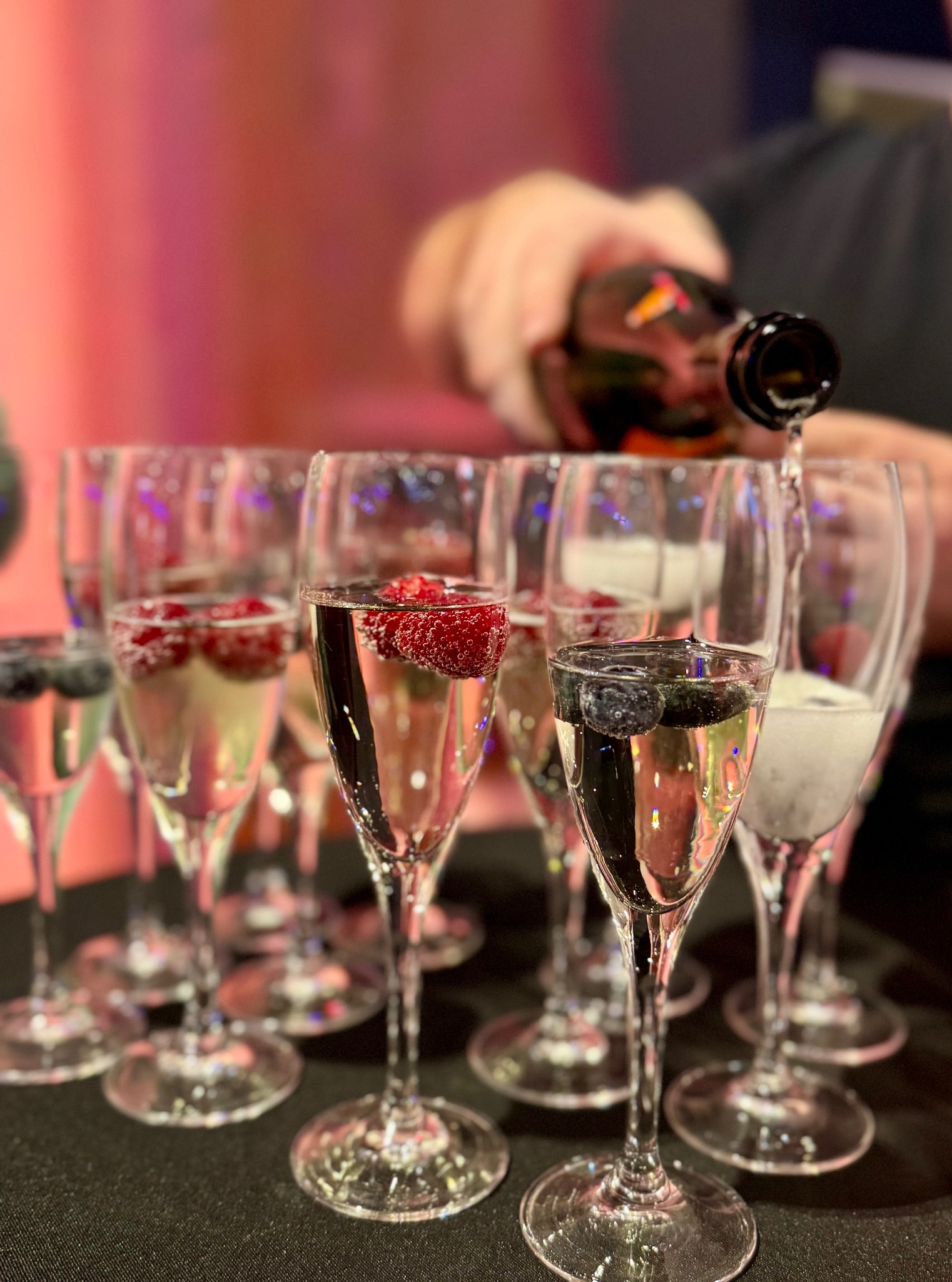 Napełnianie kieliszków szampanem z owocami (maliny, jagody) na czarnym stole, w tle różowe światło. Przygotowania do uroczystości, detale przyjęcia.