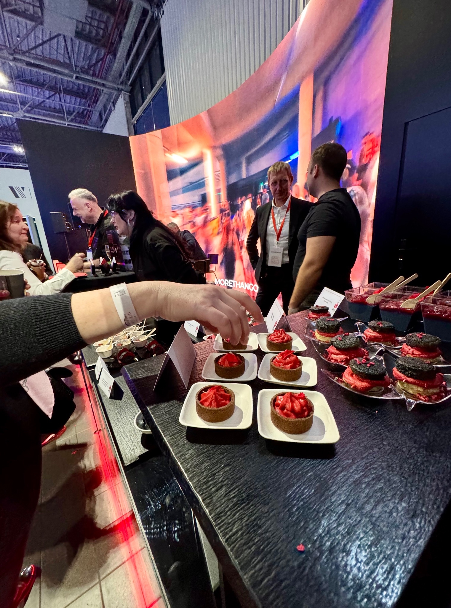 Degustacja słodkich przekąsek na evencie firmowym: tartaletki z czerwonym kremem i desery w kubeczkach. W tle osoby w strojach biznesowych i telebim.