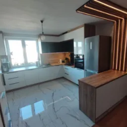 Nowoczesna kuchnia z białymi frontami, drewnianymi blatami i marmurową podłogą. Elementem wyróżniającym jest podświetlana konstrukcja z drewna nad lodówką.