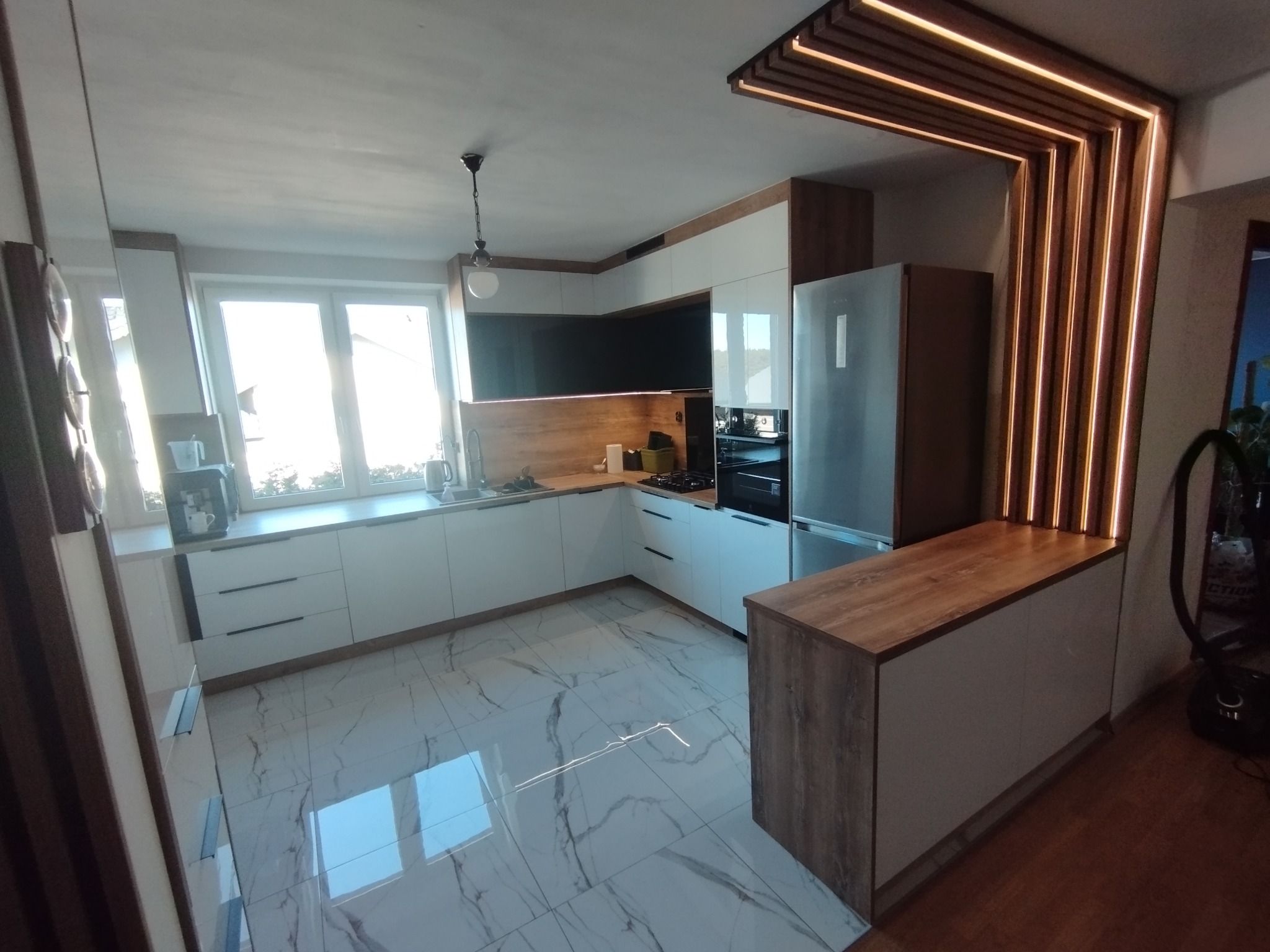 Nowoczesna kuchnia z białymi frontami, drewnianymi blatami i marmurową podłogą. Elementem wyróżniającym jest podświetlana konstrukcja z drewna nad lodówką.