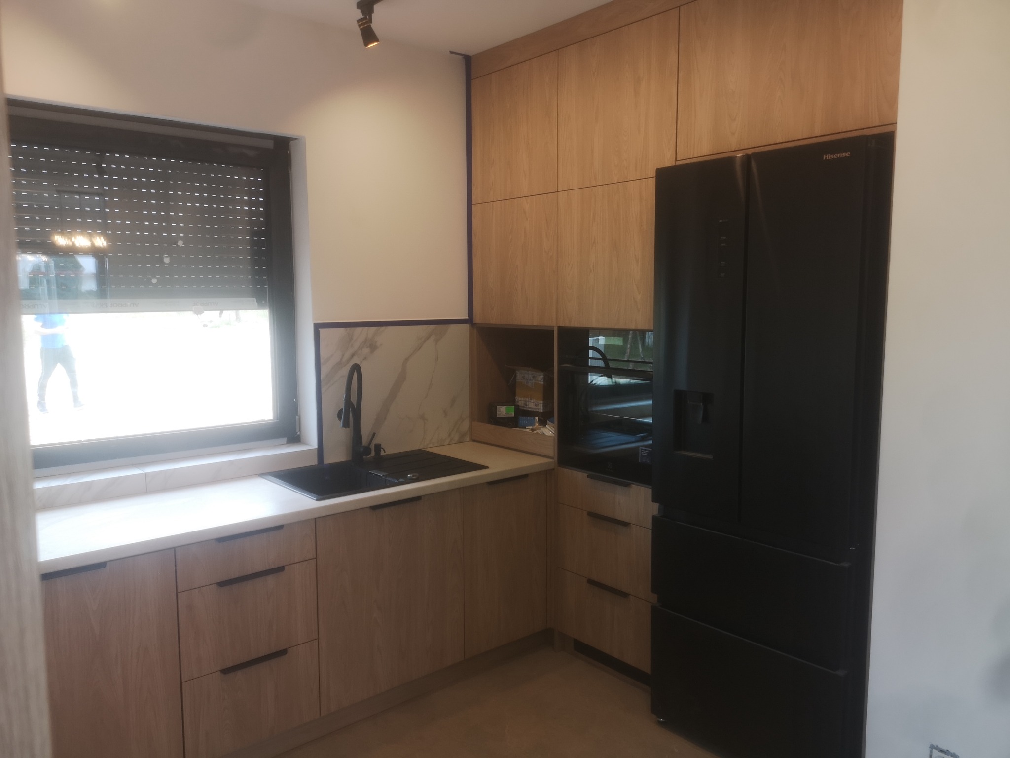 Jasna kuchnia na wymiar z drewnianymi frontami, czarnym zlewem i lodówką Hisense. Marmurowy backsplash i roleta w oknie dodają elegancji.