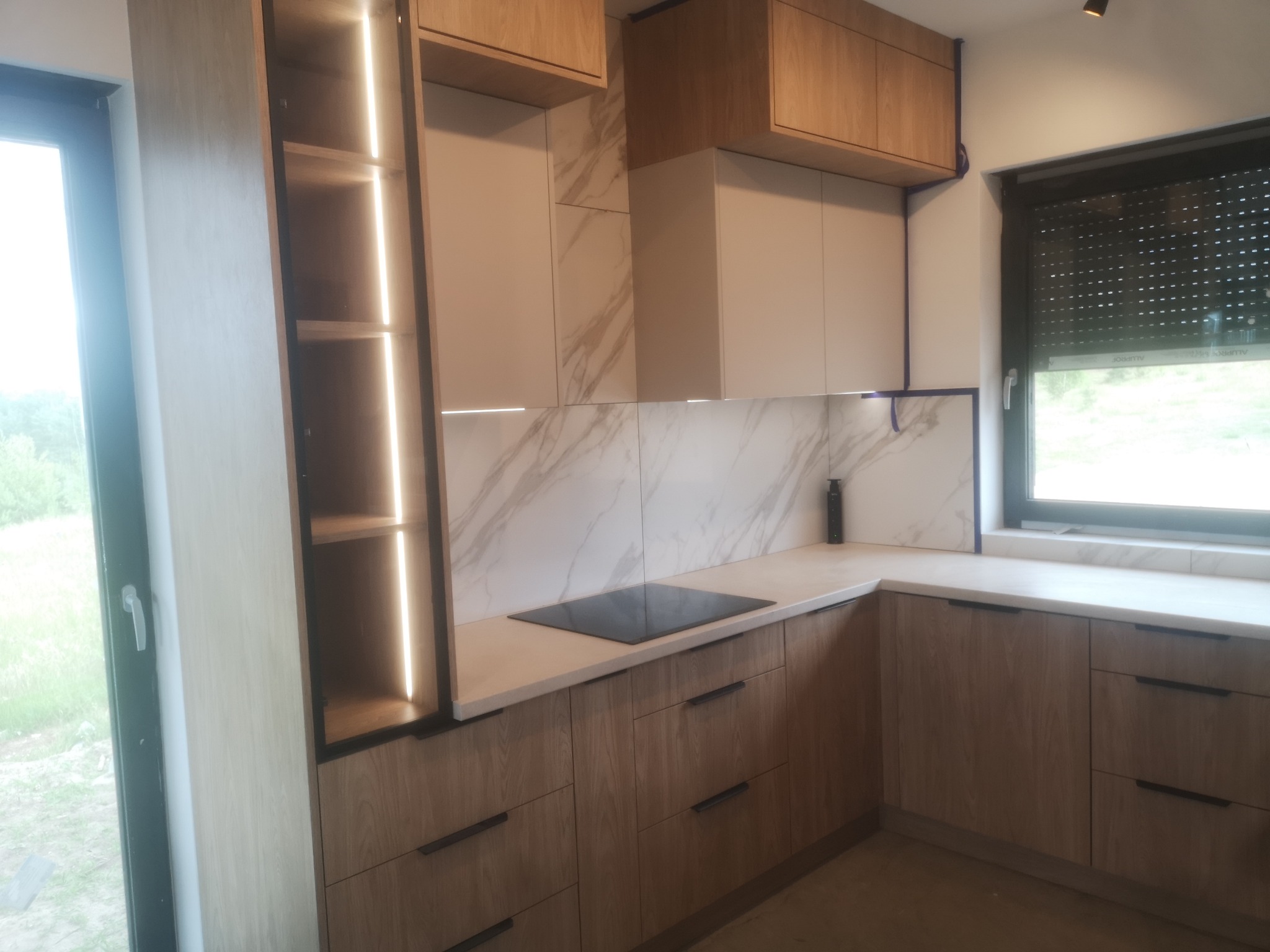 Nowoczesna kuchnia z drewnianymi frontami i marmurowym backsplashiem. Oświetlenie LED w szafce dodaje ciepła. Minimalistyczny design i funkcjonalne rozwiązania.