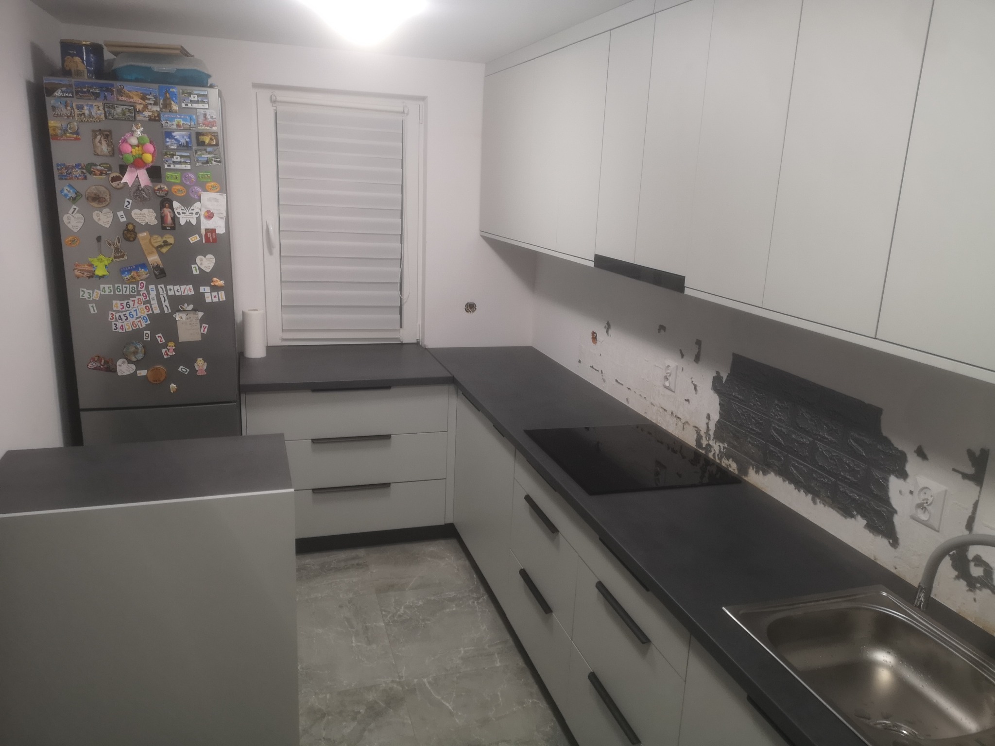 Nowoczesna kuchnia z szarymi frontami, ciemnym blatem i widocznym fragmentem ściany przed wykończeniem. Lodówka z magnesami po lewej stronie.
