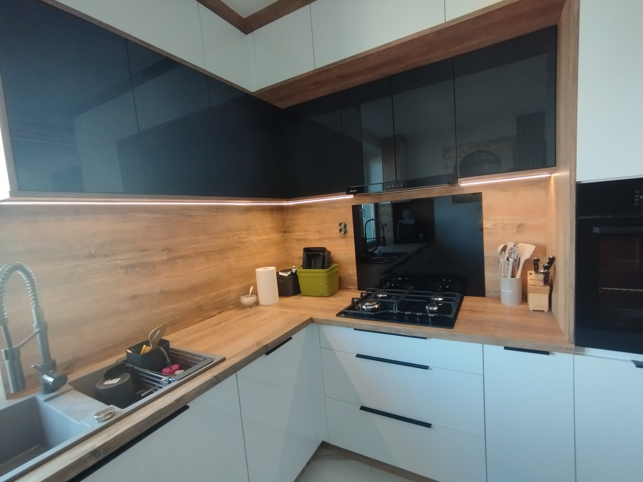 Nowoczesna kuchnia z czarnymi, przeszklonymi szafkami górnymi i białymi dolnymi. Drewniany blat i oświetlenie LED dodają ciepła. Widoczna płyta gazowa i zlewozmywak.