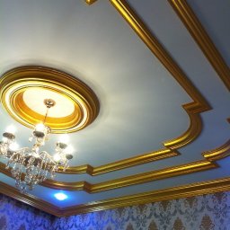 DEPOLDOM SPÓŁKA Z OGRANICZONĄ ODPOWIEDZIALNOŚCIĄ - Dekoracyjny sufit z gipsowymi zdobieniami w kolorze złota, uzupełniony kryształowym żyrandolem i wzorzystą tapetą w odcieniach błękitu i złota.