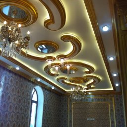 DEPOLDOM SPÓŁKA Z OGRANICZONĄ ODPOWIEDZIALNOŚCIĄ - Bogato zdobiony sufit z oświetleniem LED i kryształowymi żyrandolami w pomieszczeniu z wzorzystą tapetą i oknem łukowym. Złote detale wykończeniowe.