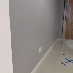 DEPOLDOM SPÓŁKA Z OGRANICZONĄ ODPOWIEDZIALNOŚCIĄ - Świeżo pomalowana na szaro ściana z białą listwą przypodłogową i gniazdkiem elektrycznym. Podłoga wyłożona jasnymi płytkami. Drzwi zabezpieczone folią malarską.