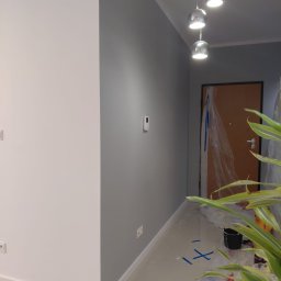 DEPOLDOM SPÓŁKA Z OGRANICZONĄ ODPOWIEDZIALNOŚCIĄ - Świeżo pomalowane na szaro ściany w korytarzu, widoczne zabezpieczenia podłogi folią malarską. W tle drzwi wejściowe i wiszące lampy.