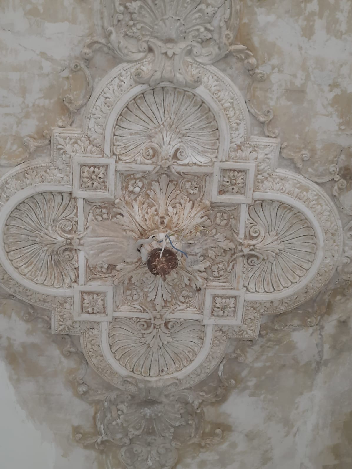 Dekoracyjny, kremowy sufit z gipsowymi zdobieniami w stylu retro. W centrum widoczny zaczep na lampę. Widoczne niedoskonałości i ślady czasu.