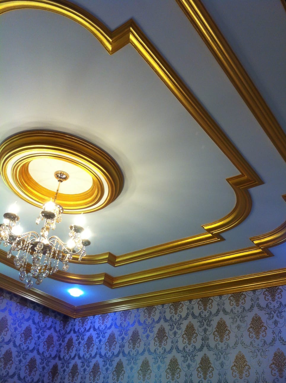 Dekoracyjny sufit z gipsowymi zdobieniami w kolorze złota, uzupełniony kryształowym żyrandolem i wzorzystą tapetą w odcieniach błękitu i złota.
