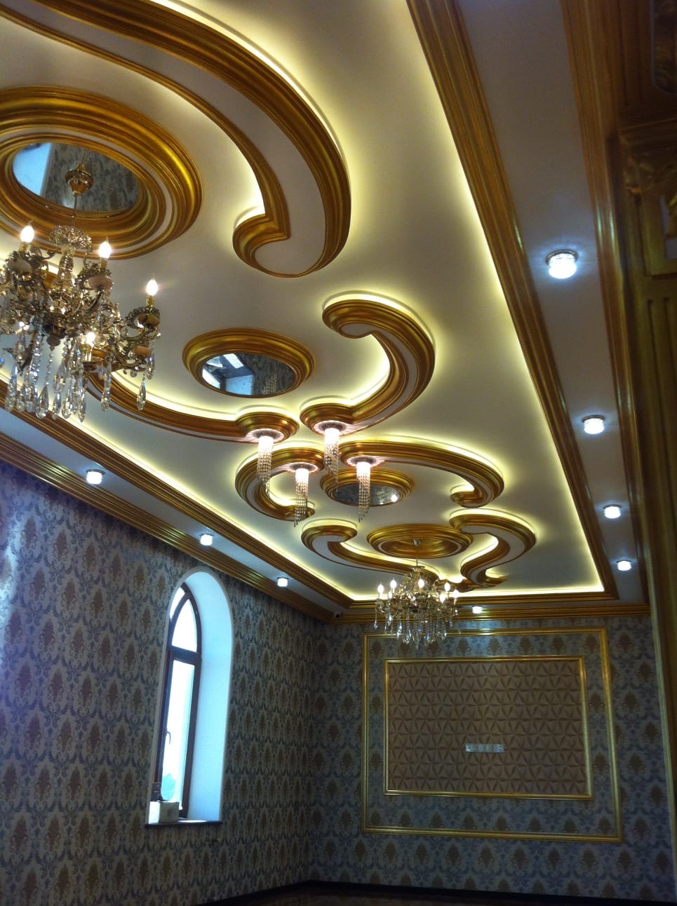 Bogato zdobiony sufit z oświetleniem LED i kryształowymi żyrandolami w pomieszczeniu z wzorzystą tapetą i oknem łukowym. Złote detale wykończeniowe.