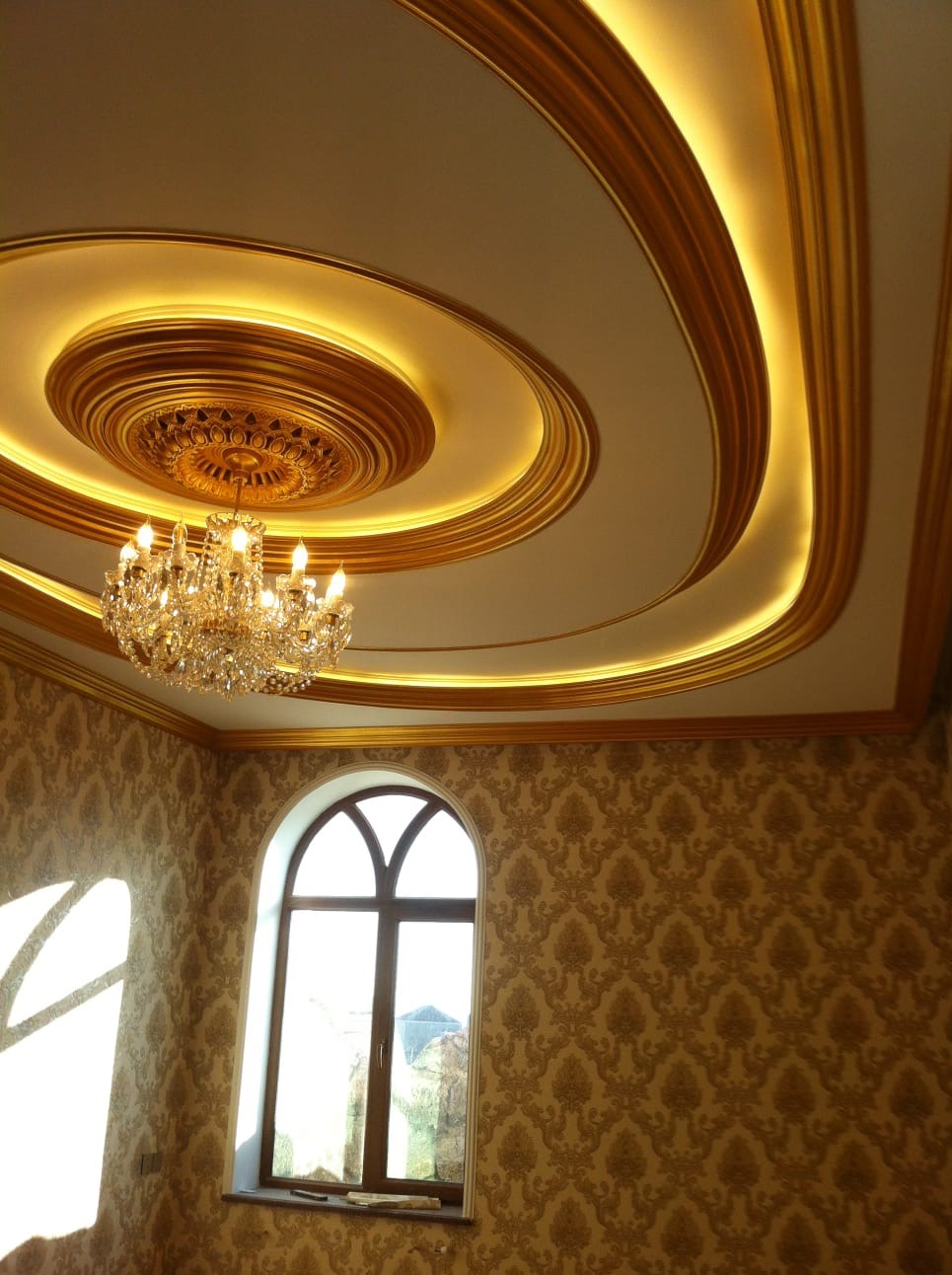 Dekoracyjny sufit z oświetleniem LED i żyrandolem kryształowym, ściany z wzorzystą tapetą i łukowe okno, tworzące eleganckie wykończenie wnętrza.
