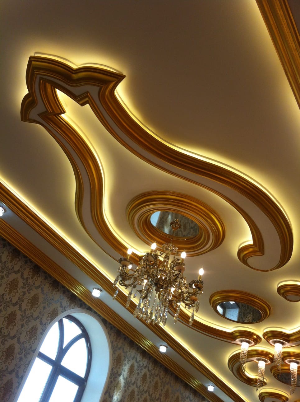 Dekoracyjny sufit z podświetleniem LED i złoceniami, widok z dołu. Kryształowy żyrandol w centrum. Bogato zdobione wnętrze z oknem łukowym w tle.