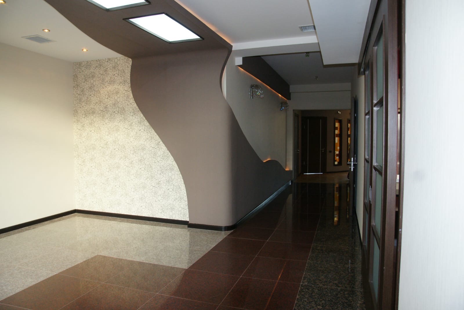 Nowoczesny korytarz z dekoracyjną ścianą w odcieniach beżu i brązu, wzorzysta tapeta, błyszcząca podłoga z płytek. Oświetlenie sufitowe podkreśla unikalny design wnętrza.