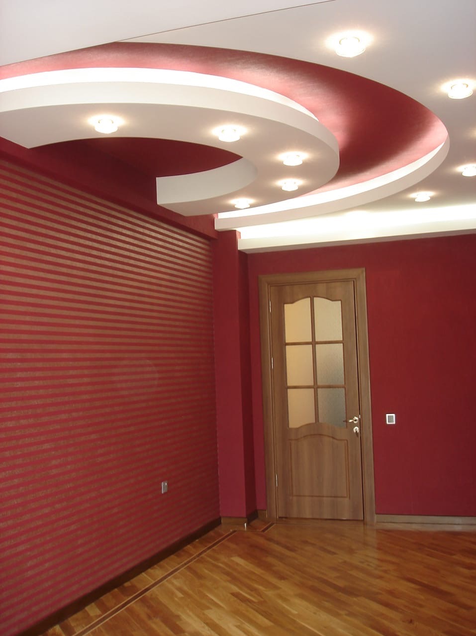 Czerwone wnętrze z dekoracyjnym sufitem podwieszanym, oświetleniem punktowym i drewnianą podłogą. Paski na ścianie dodają tekstury. Drzwi z szybami w tle.