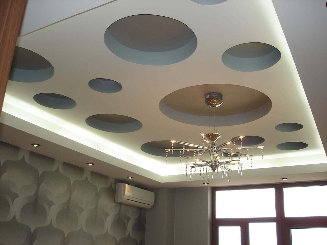 Dekoracyjny sufit podwieszany z okrągłymi wnękami i oświetleniem LED, centralnie umieszczony żyrandol. W tle widoczna ściana z wzorzystą tapetą i klimatyzator.
