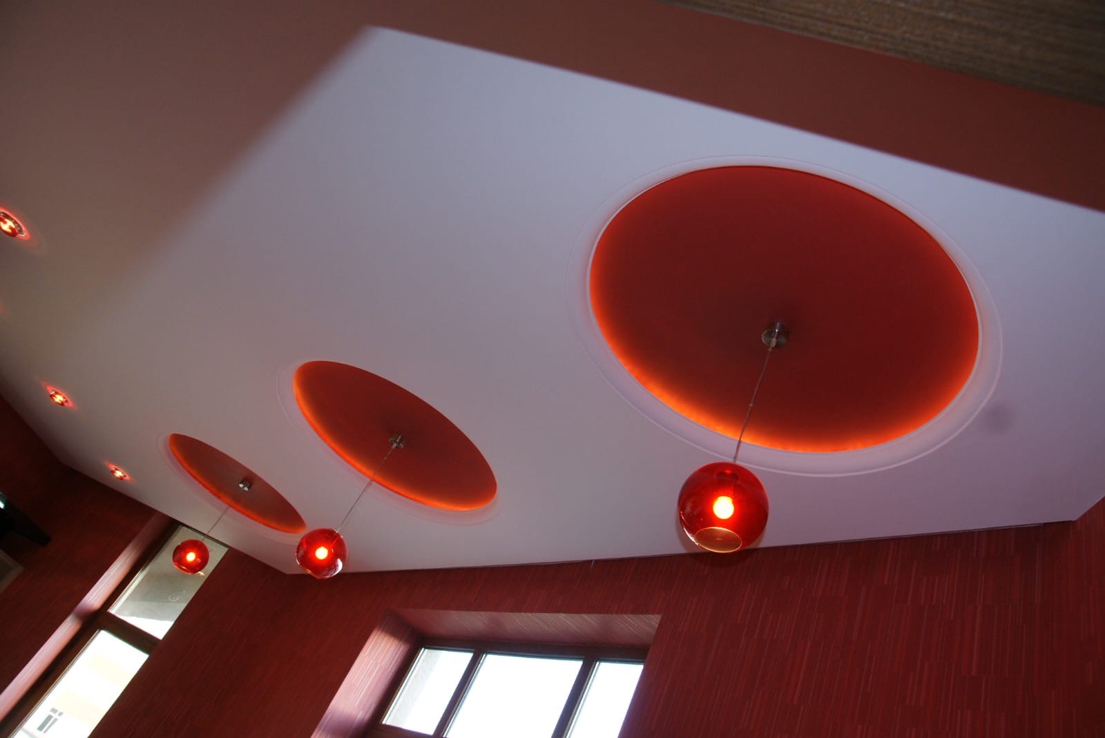 Nowoczesny sufit podwieszany z okrągłymi wnękami podświetlonymi na pomarańczowo i szklanymi, kulistymi lampami wiszącymi w pokoju z czerwonymi ścianami i oknami.