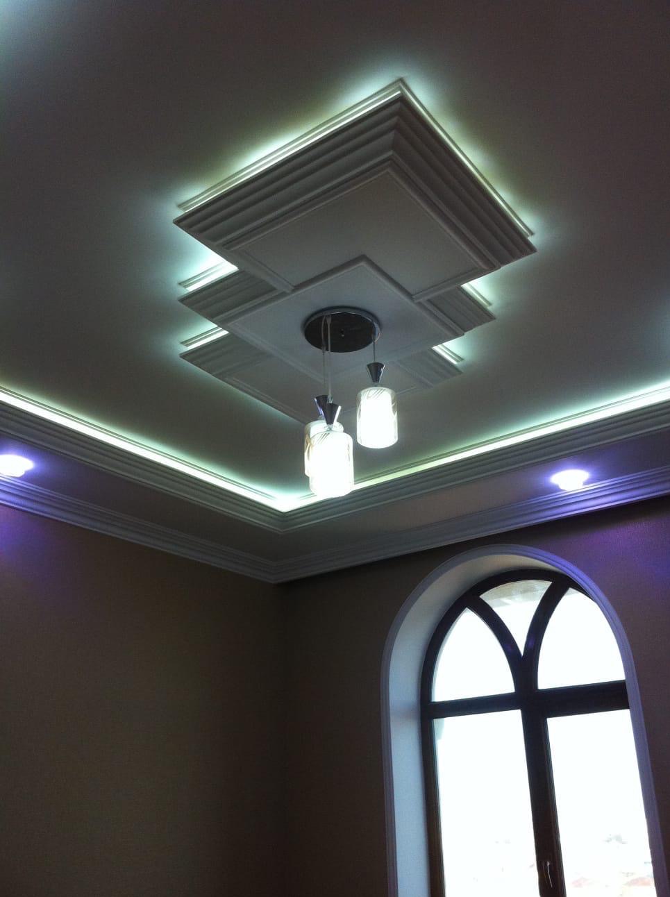 Dekoracyjny sufit z oświetleniem LED i lampami wiszącymi, widok z dołu, fragment okna łukowego. Aranżacja wnętrza w jasnych kolorach, nowoczesny design.