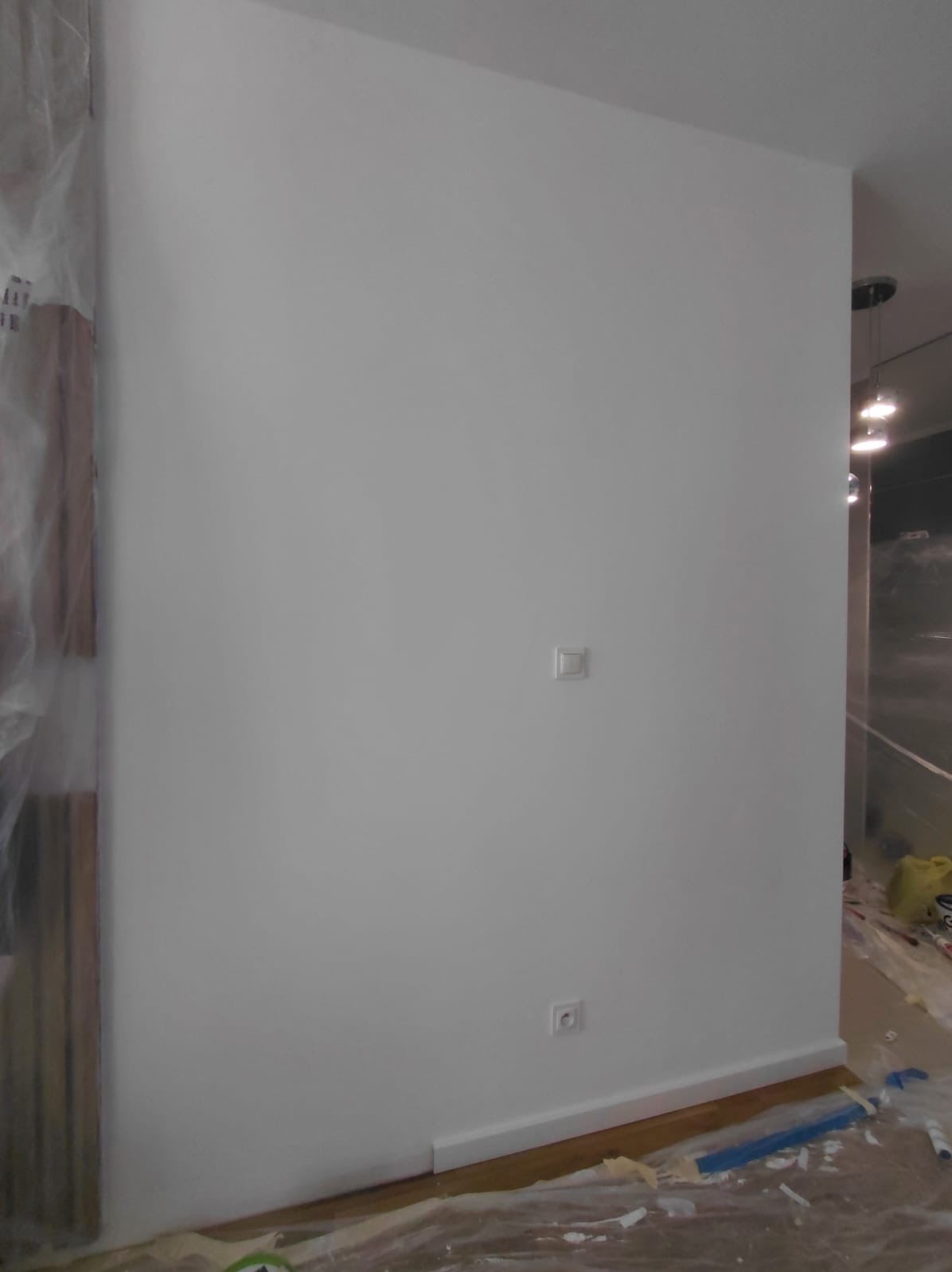 Świeżo pomalowana na biało ściana z gładkimi powierzchniami, widoczne gniazdka elektryczne, listwa przypodłogowa i folia ochronna na podłodze w trakcie prac wykończeniowych.