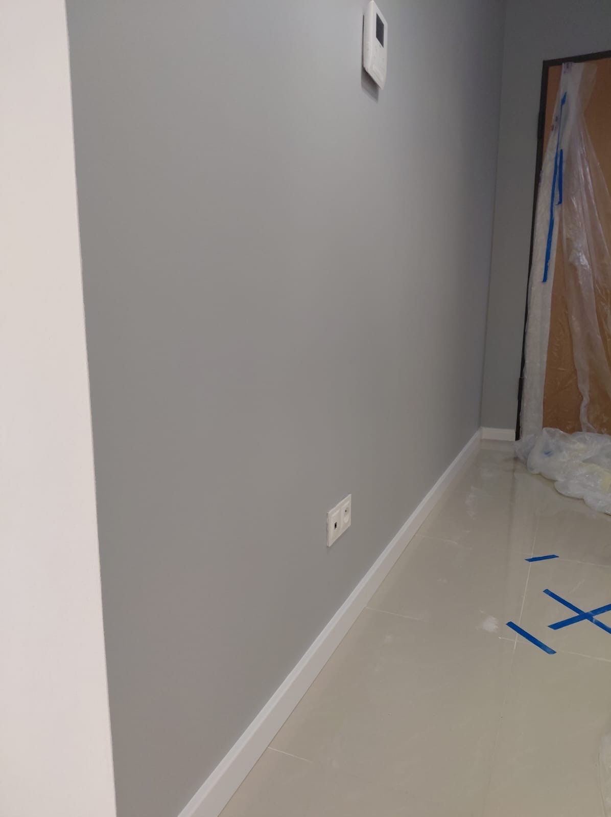 Świeżo pomalowana na szaro ściana z białą listwą przypodłogową i gniazdkiem elektrycznym. Podłoga wyłożona jasnymi płytkami. Drzwi zabezpieczone folią malarską.