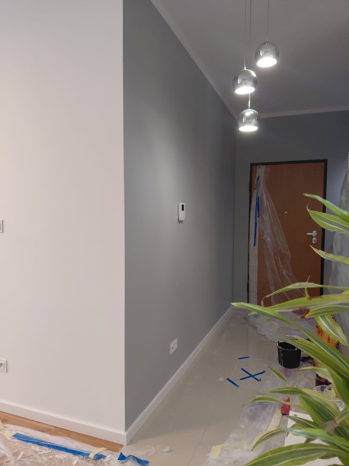 Świeżo pomalowane na szaro ściany w korytarzu, widoczne zabezpieczenia podłogi folią malarską. W tle drzwi wejściowe i wiszące lampy.