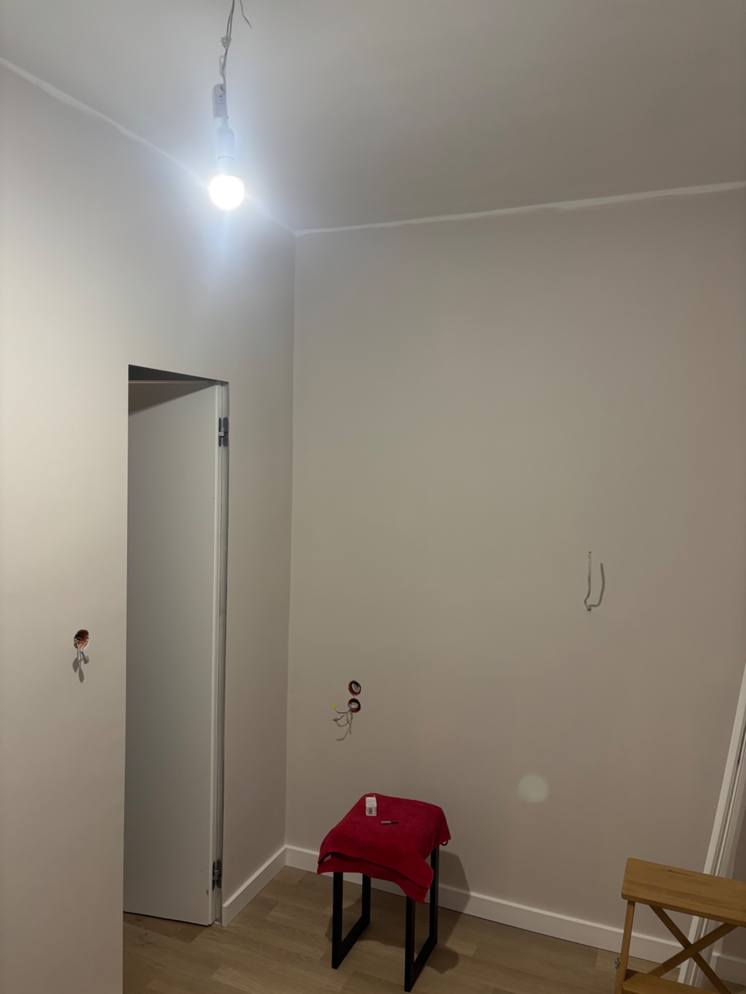 Wnętrze pokoju w trakcie remontu: pomalowane ściany w odcieniach beżu, widoczne kable, listwy przypodłogowe, drabina malarska i stolik z czerwonym materiałem.