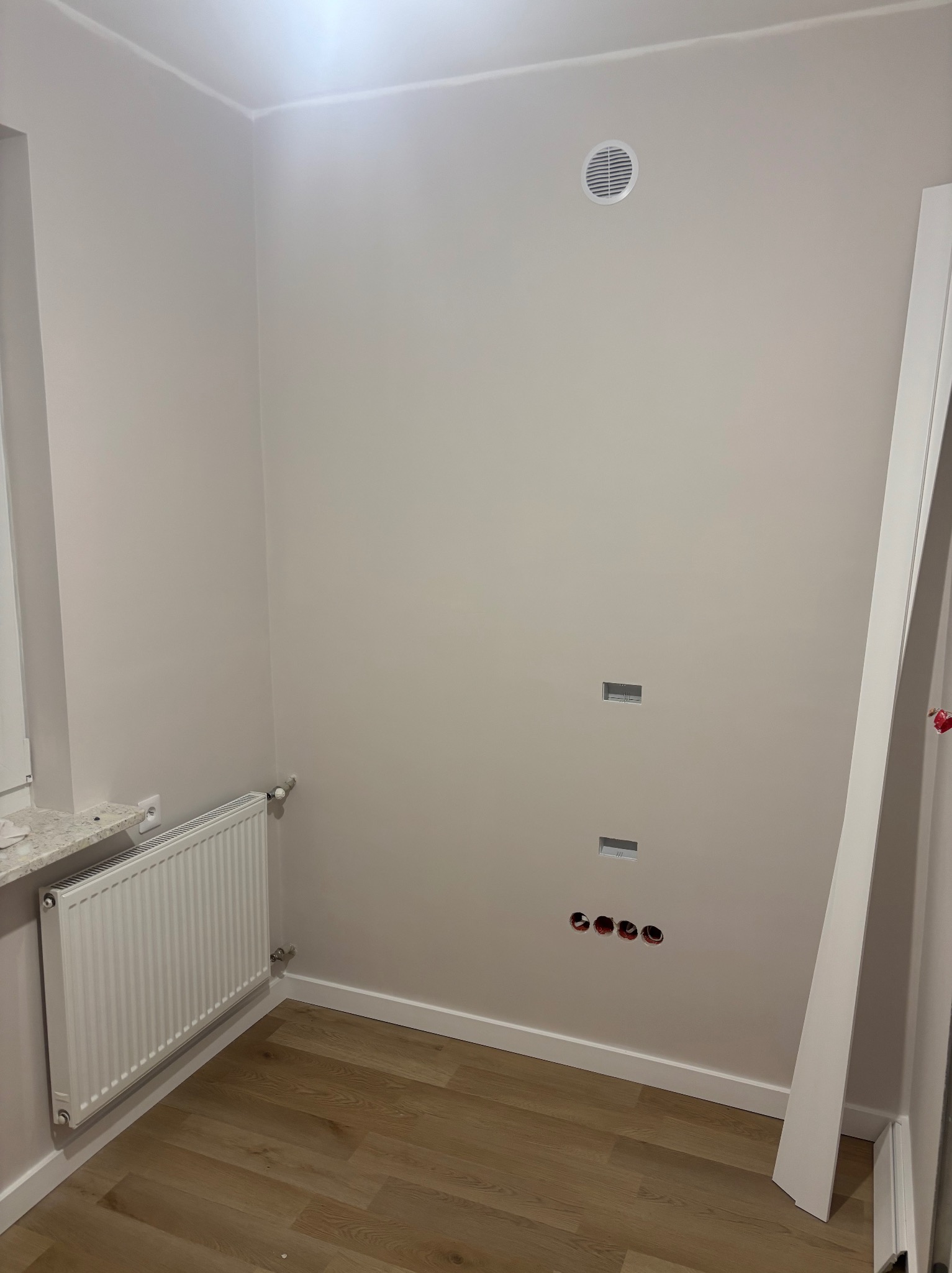 Jasne wnętrze w trakcie wykańczania: pomalowane ściany, grzejnik, listwa przypodłogowa, otwory na gniazdka. Widoczny fragment podłogi z paneli.