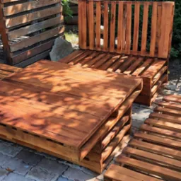 Komplet mebli ogrodowych z palet: stół i sofy, pomalowane na brązowo, ustawione na kostce brukowej. W tle zieleń ogrodu i grill.