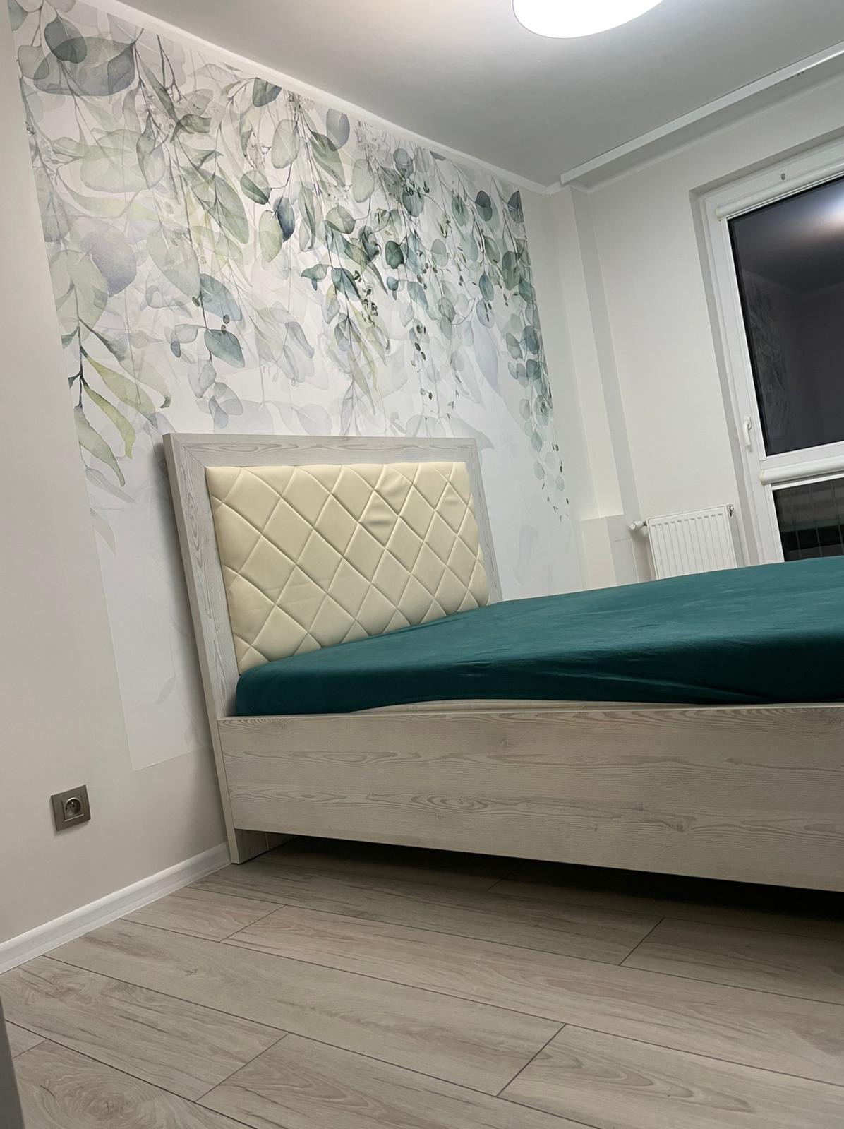 Białe łóżko z pikowanym zagłówkiem i zieloną pościelą na tle ściany z motywem roślinnym. Jasna podłoga z paneli. Widok na gniazdko elektryczne i okno.