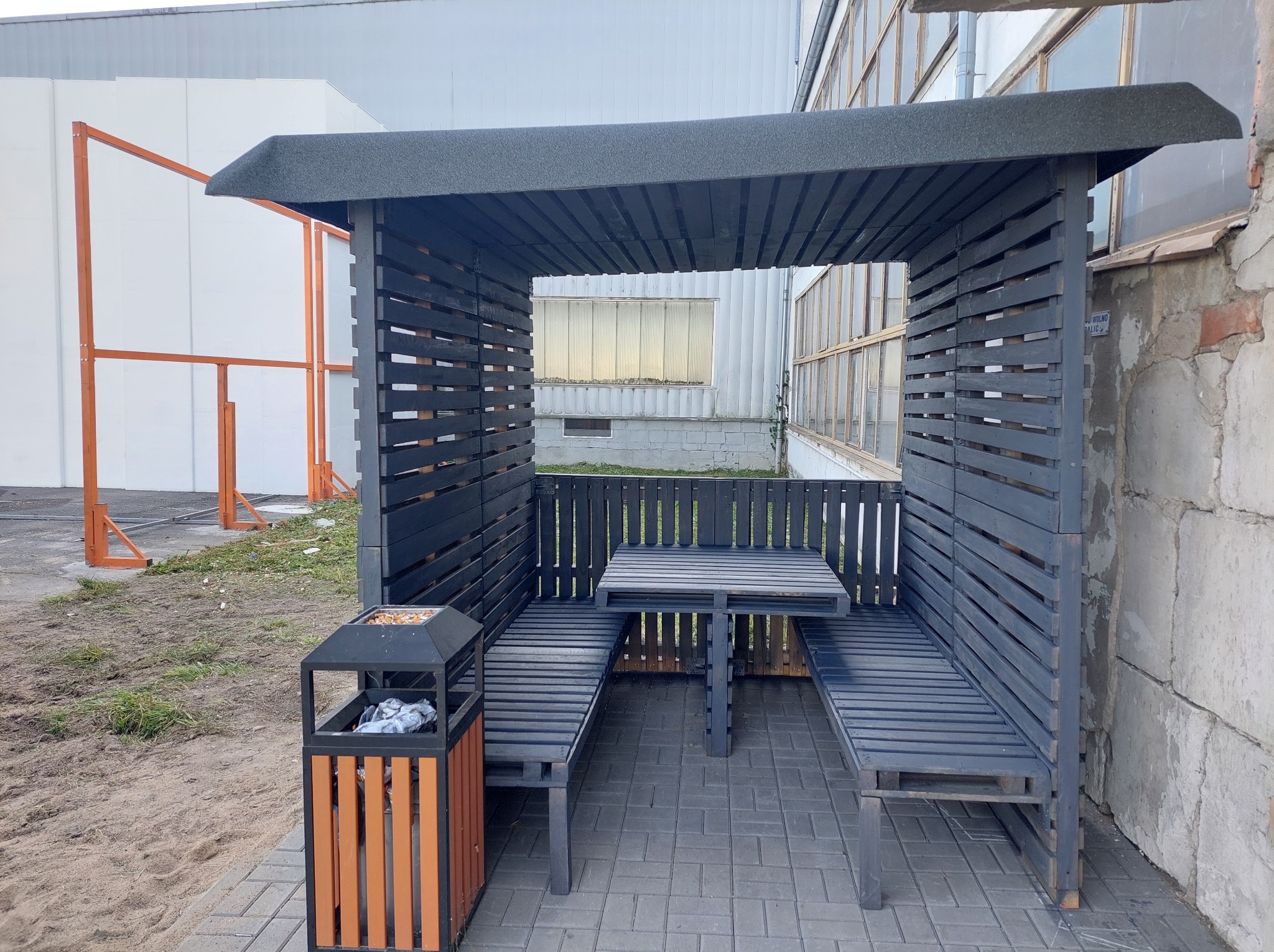 Drewniana altana pomalowana na szaro z ławkami i stołem w środku. Obok stoi kosz na śmieci. Na zewnątrz budynek przemysłowy z oknami i fragment konstrukcji pomalowanej na pomarańczowo.