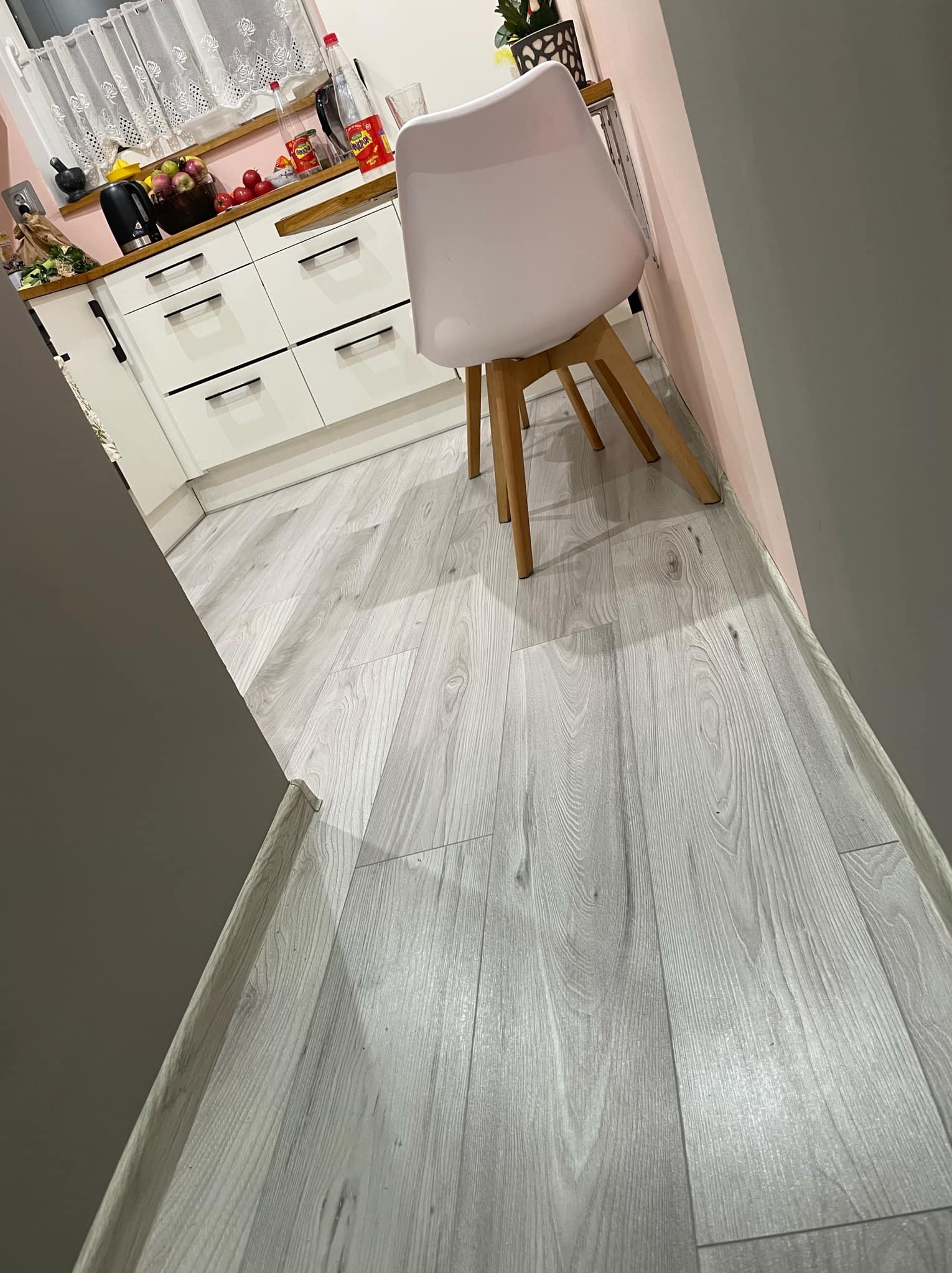 Jasnoszara podłoga w kuchni z białymi meblami i krzesłem. Widok z perspektywy wejścia, z fragmentem ściany po lewej stronie.