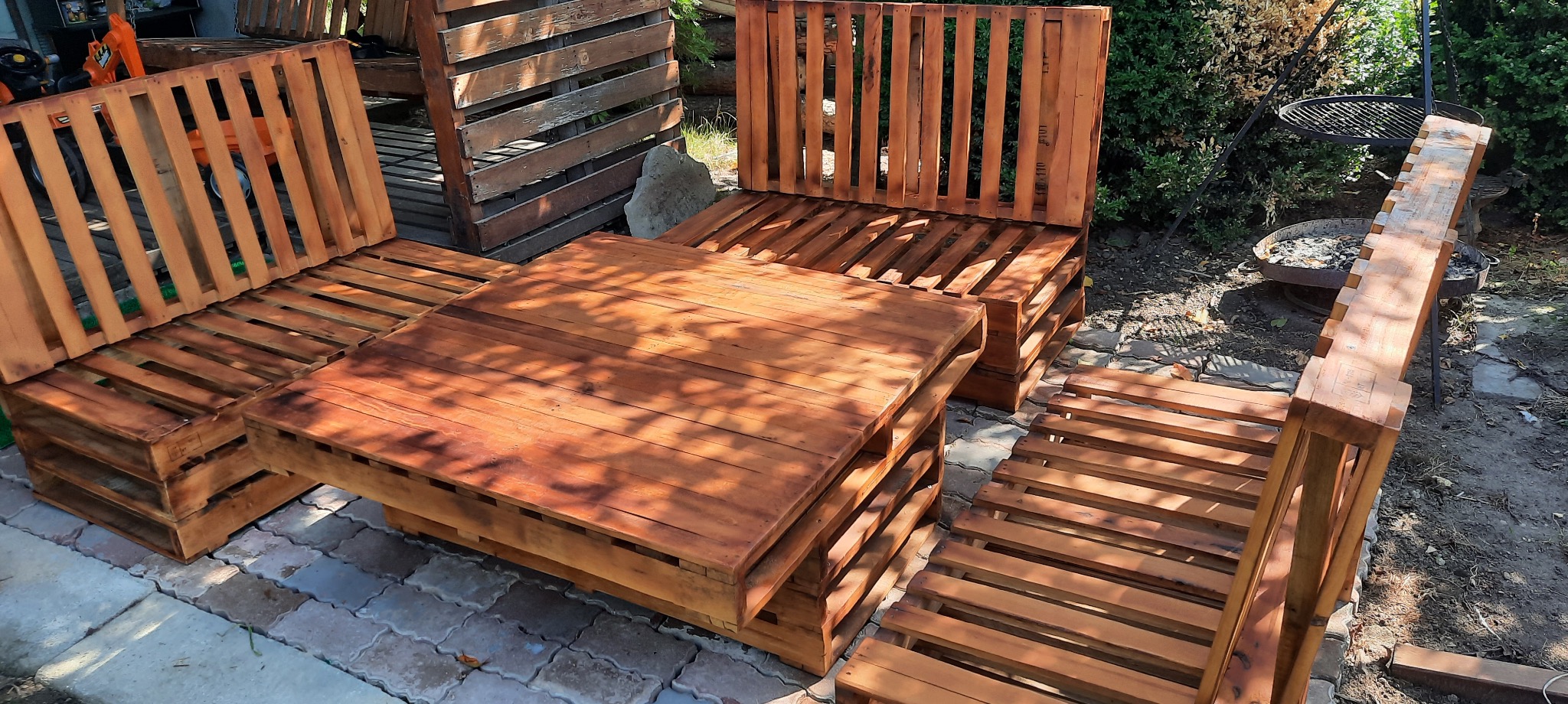 Komplet mebli ogrodowych z palet: stół i sofy, pomalowane na brązowo, ustawione na kostce brukowej. W tle zieleń ogrodu i grill.