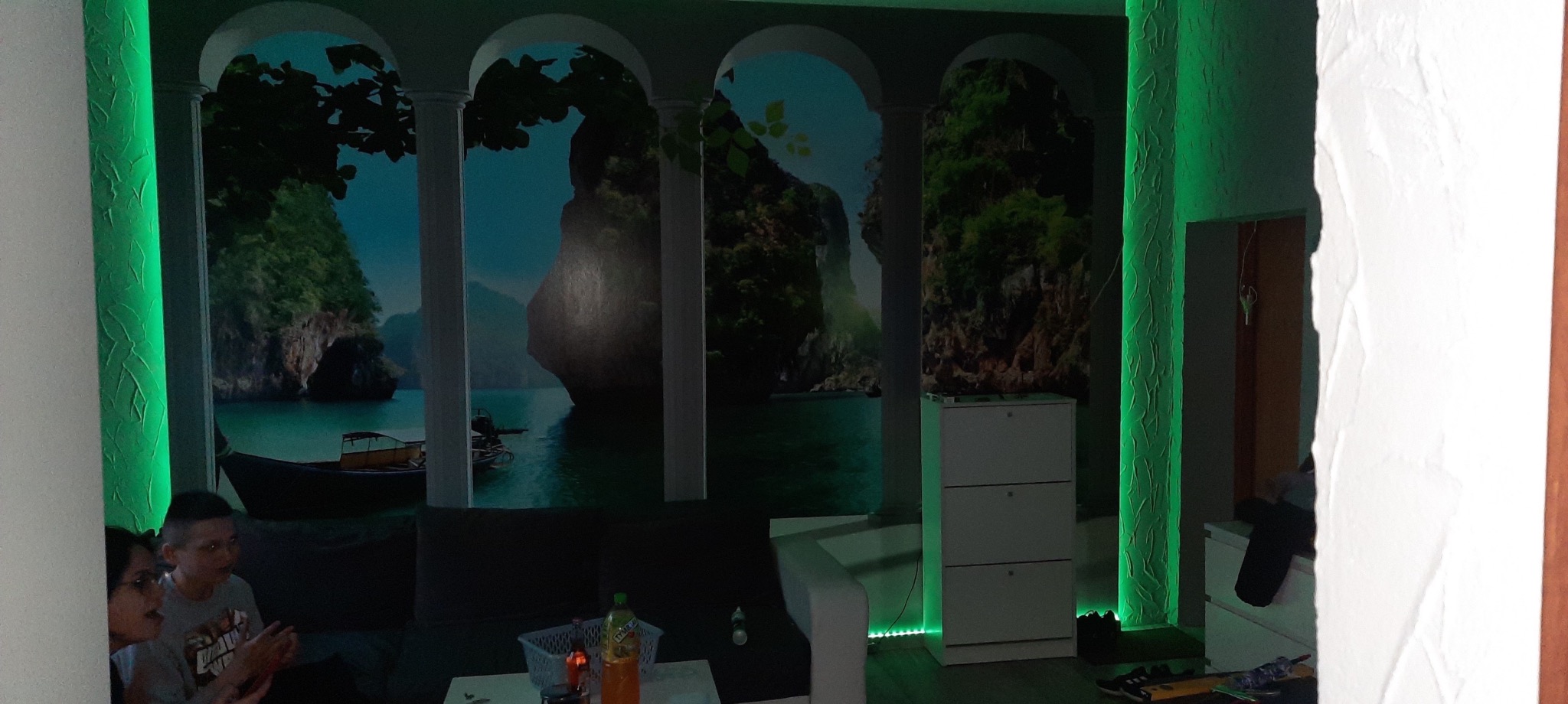 Salon z fototapetą imitującą widok na morze przez arkady, podświetlony zielonymi ledami, białe meble i osoby w tle.