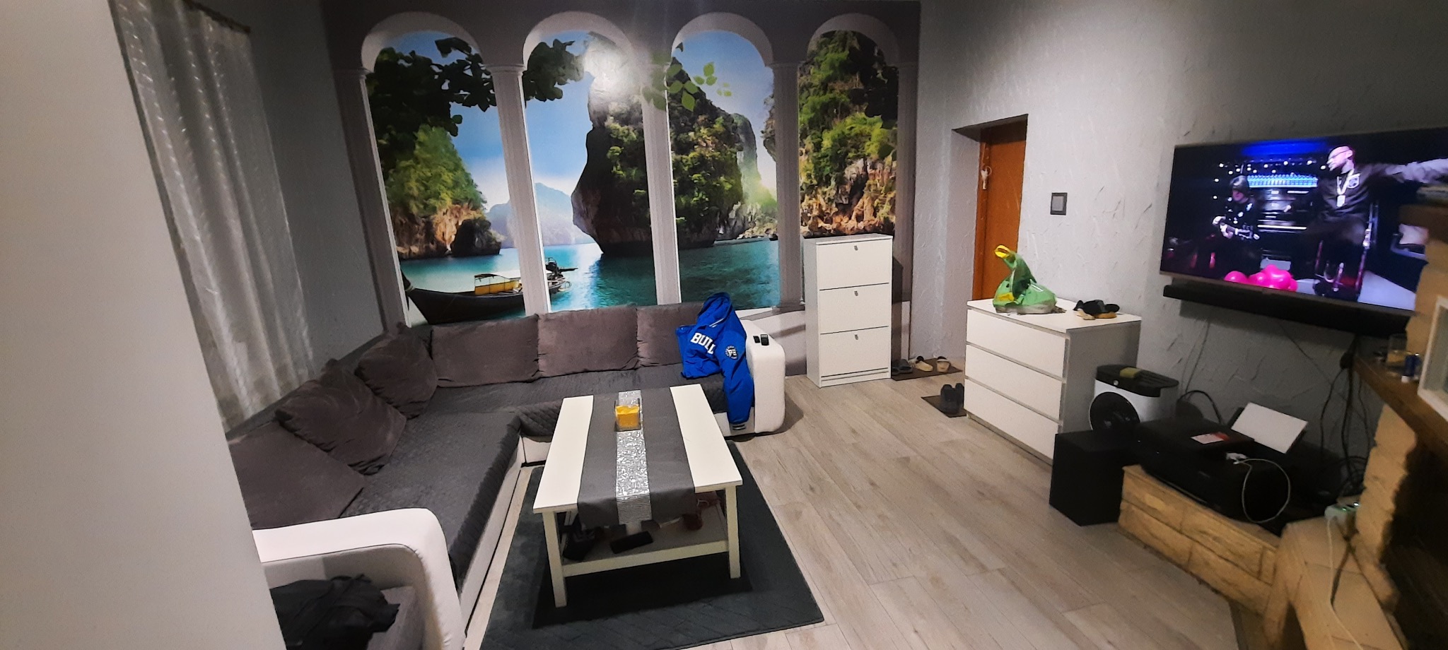 Jasne wnętrze pokoju z szarą sofą narożną, białym stolikiem kawowym i fototapetą z widokiem na morze. Na podłodze panele, a na ścianie telewizor.