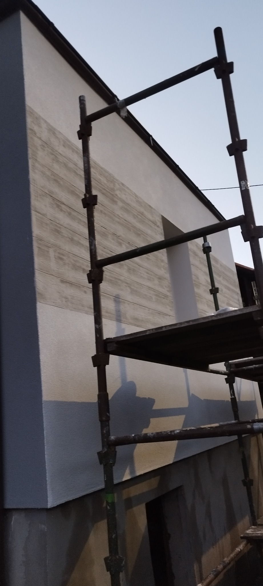 Ściana budynku w trakcie prac elewacyjnych, widoczne rusztowanie i fragmenty nałożonego tynku strukturalnego imitującego drewno. Częściowo pomalowana ściana.