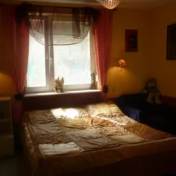 Pokój z łóżkiem w patchworkowej narzucie, zasłonięte okno z figurką anioła na parapecie i pluszowy miś na kanapie w tle.