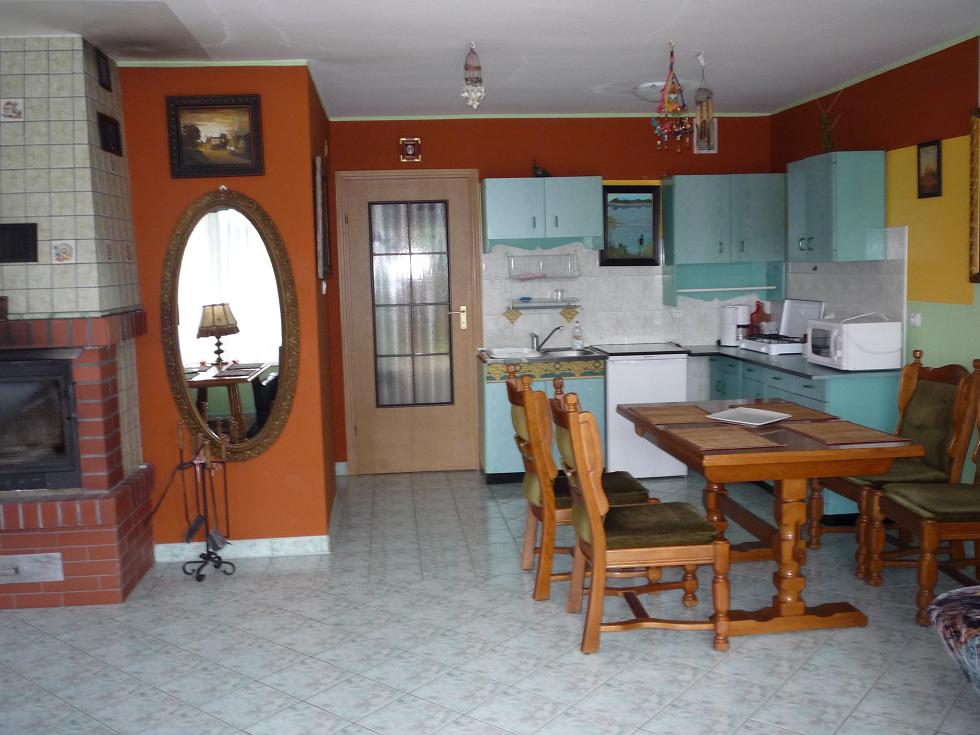 Wnętrze pokoju z kominkiem, kuchnią w tle i drewnianym stołem z krzesłami, odbicie lampy w ozdobnym lustrze.