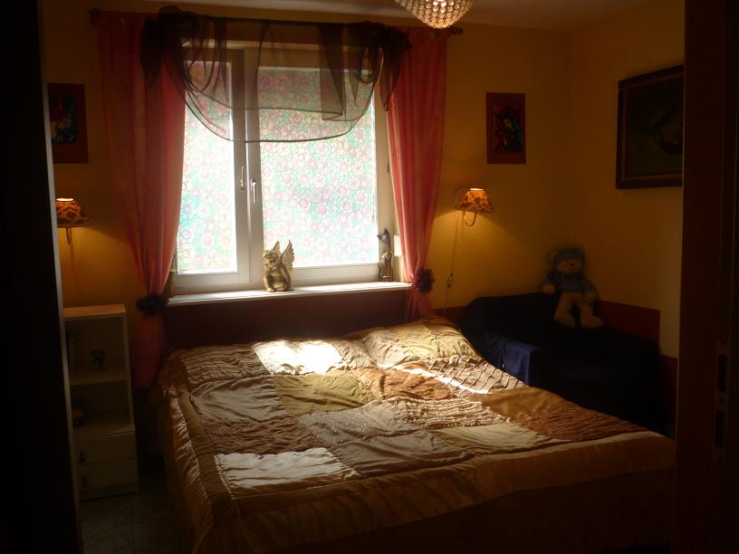 Pokój z łóżkiem w patchworkowej narzucie, zasłonięte okno z figurką anioła na parapecie i pluszowy miś na kanapie w tle.