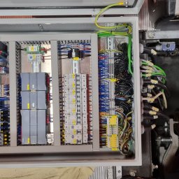 IA - INDUSTRIAL AUTOMATION SP. Z O.O. - Otwarta szafa sterownicza z modułami PLC, bezpiecznikami i okablowaniem. Widoczne detale montażu i podłączeń. Elementy automatyki przemysłowej.