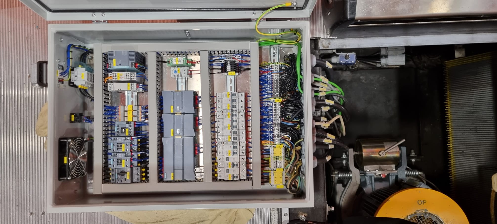 Otwarta szafa sterownicza z modułami PLC, bezpiecznikami i okablowaniem. Widoczne detale montażu i podłączeń. Elementy automatyki przemysłowej.