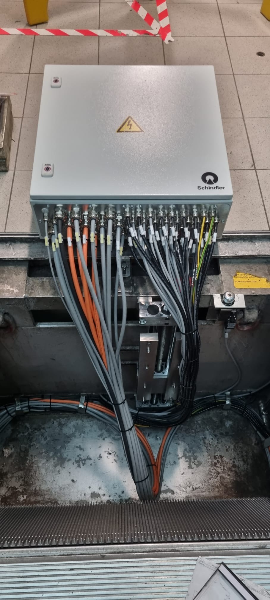 Szara skrzynka elektryczna Schindler z licznymi kablami w kolorach szarym, pomarańczowym i czarnym, podłączonymi do złączek. Widoczny znak ostrzegawczy.