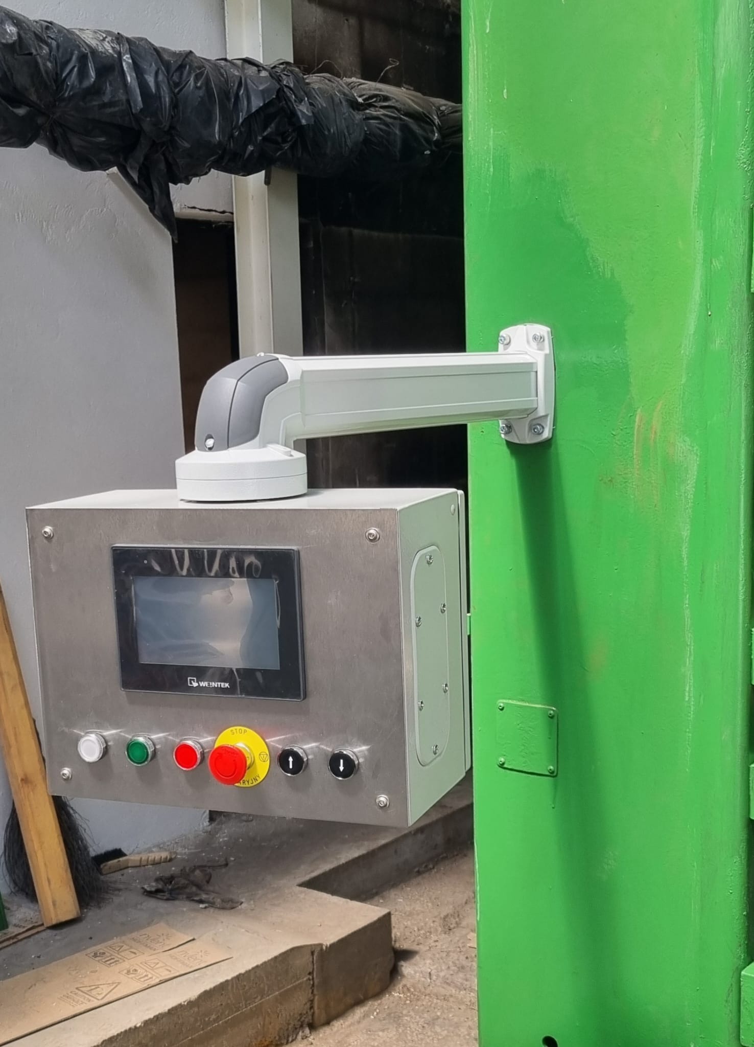 Panel sterowania z ekranem dotykowym i przyciskami zamontowany na zielonej ścianie, z widocznym okablowaniem i elementami konstrukcyjnymi.