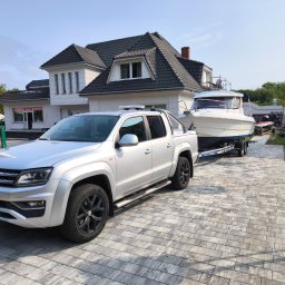 GRZEGORZ ZALESKI POMOC DROGOWA KWADRAT - Srebrny pick-up Volkswagen Amarok z przyczepą i łodzią na posesji z kostki brukowej. Widoczny budynek w tle. Transport łodzi.