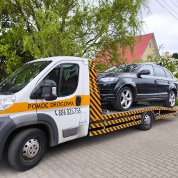 GRZEGORZ ZALESKI POMOC DROGOWA KWADRAT - Laweta z czarnym SUV-em Audi Q7 na platformie, zaparkowana na brukowanej drodze, z zielenią w tle. Na lawecie napis 'Pomoc Drogowa' i numer telefonu.