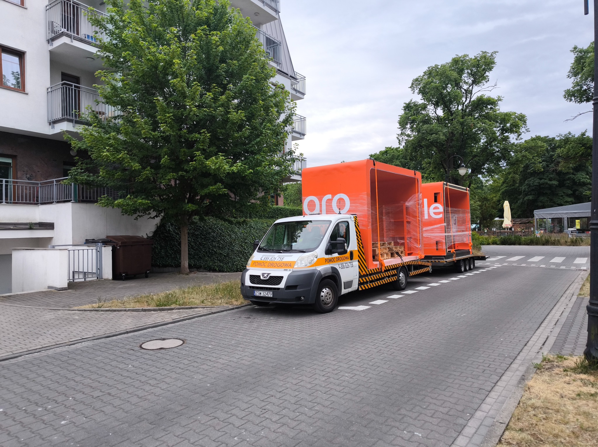 Pomoc drogowa na lawecie z pomarańczową zabudową, zaparkowana na ulicy osiedlowej z budynkiem mieszkalnym w tle. Na zabudowie widoczne logo.