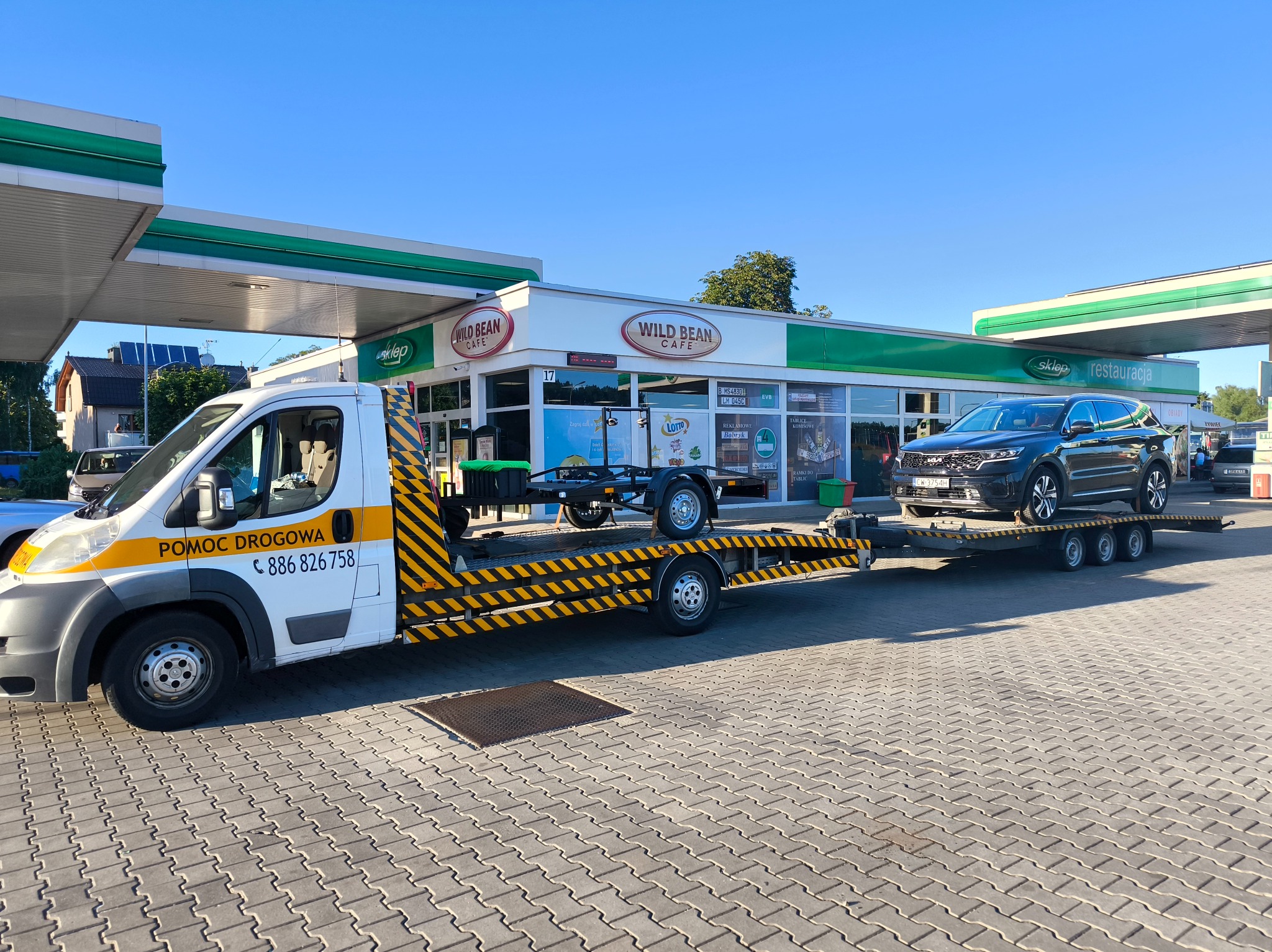 Laweta z czarno-żółtymi pasami przewozi czarny samochód osobowy na tle stacji benzynowej Wild Bean Cafe w słoneczny dzień. Widoczny numer telefonu pomocy drogowej.