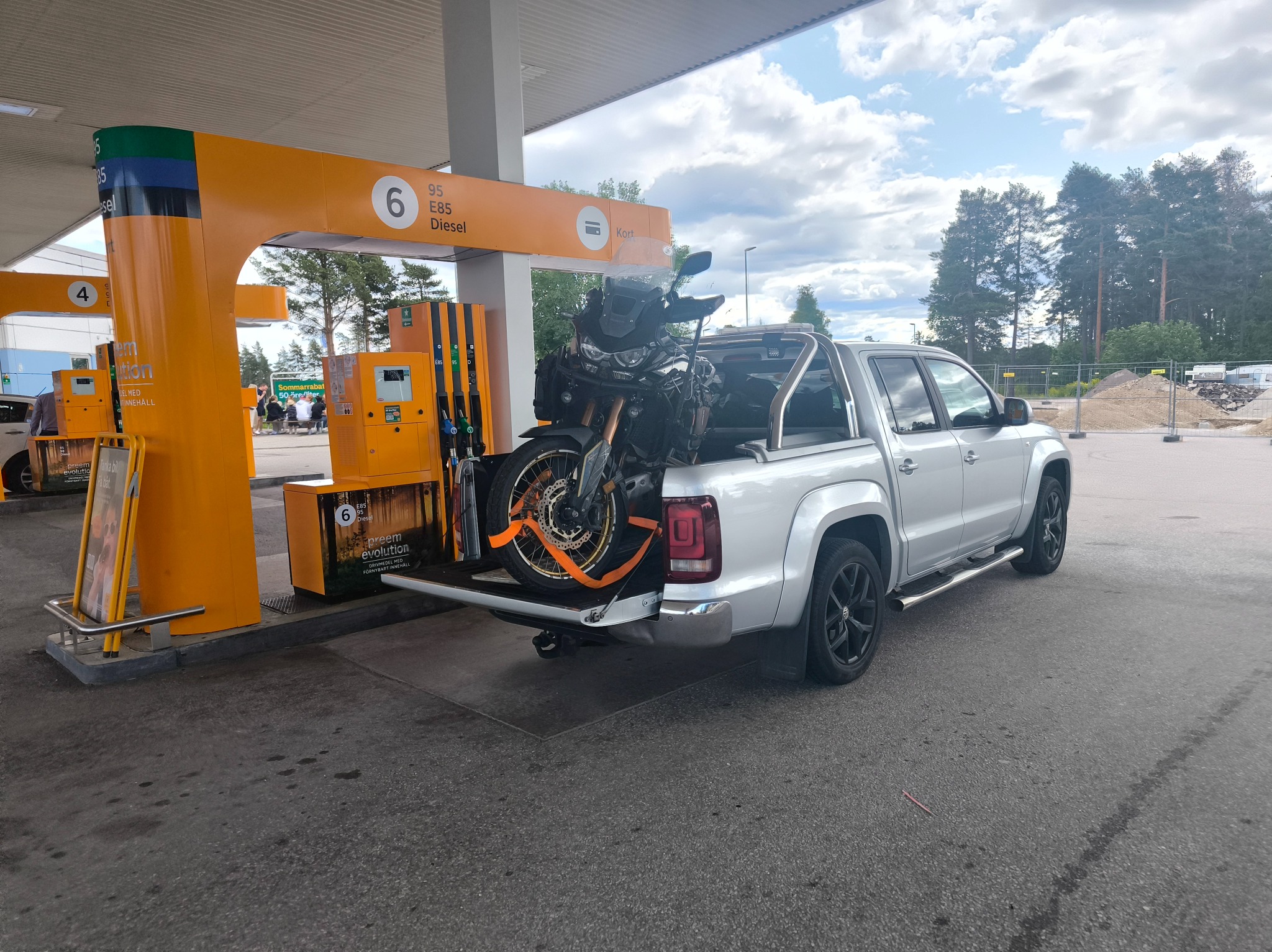 Srebrny pickup z motocyklem na pace, zabezpieczonym pomarańczowymi pasami, stoi na stacji paliw Preem. W tle dystrybutory i zieleń.