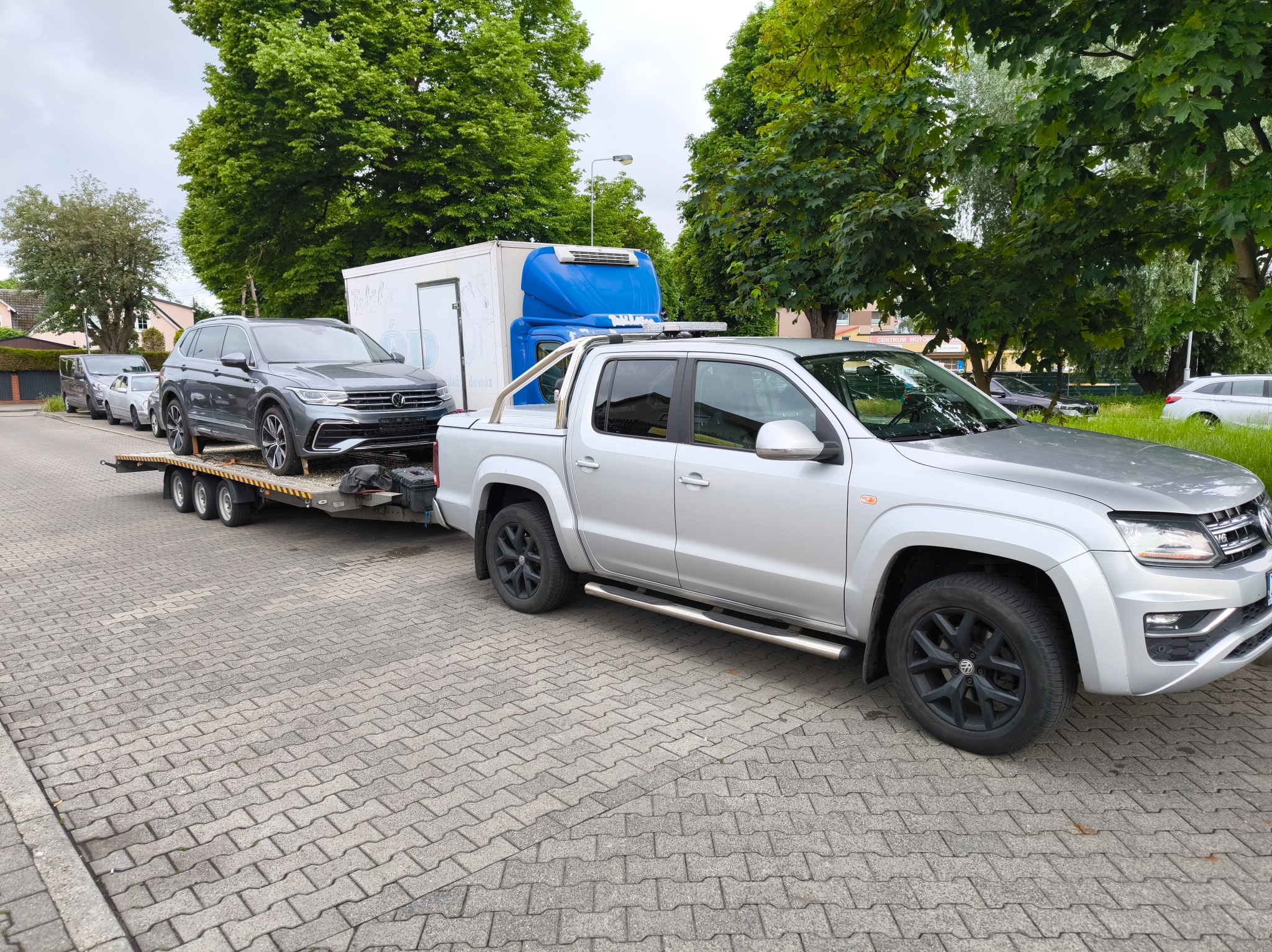 Laweta z szarym Volkswagenem Tiguan na przyczepie, srebrny pickup Volkswagen Amarok z kabiną załadowczą, drzewa w tle, kostka brukowa.